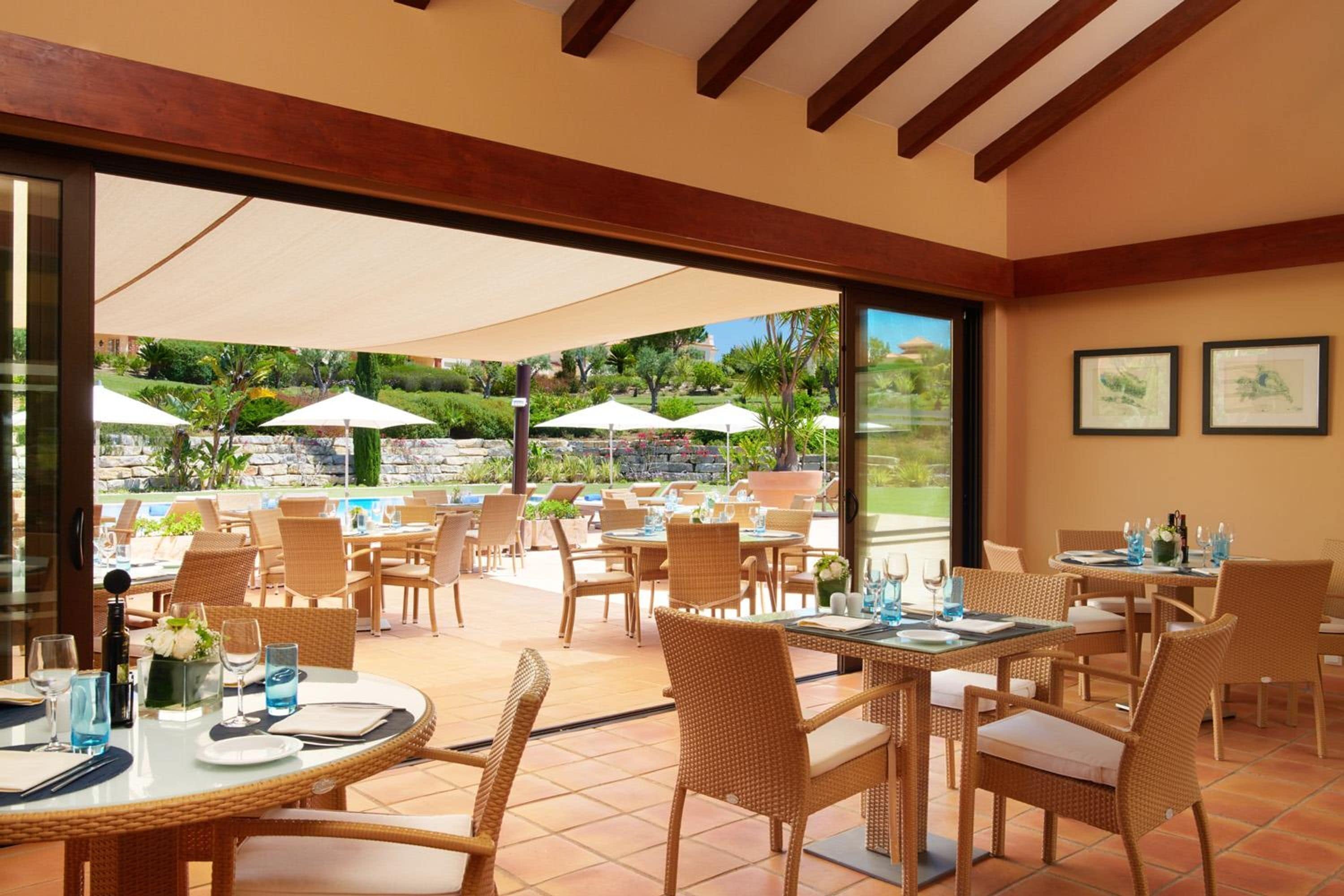 Un área de comedor al aire libre con sillas y mesas de mimbre, sombrillas grandes y vistas a una piscina y follaje.