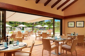 Un área de comedor al aire libre con sillas y mesas de mimbre, sombrillas grandes y vistas a una piscina y follaje.