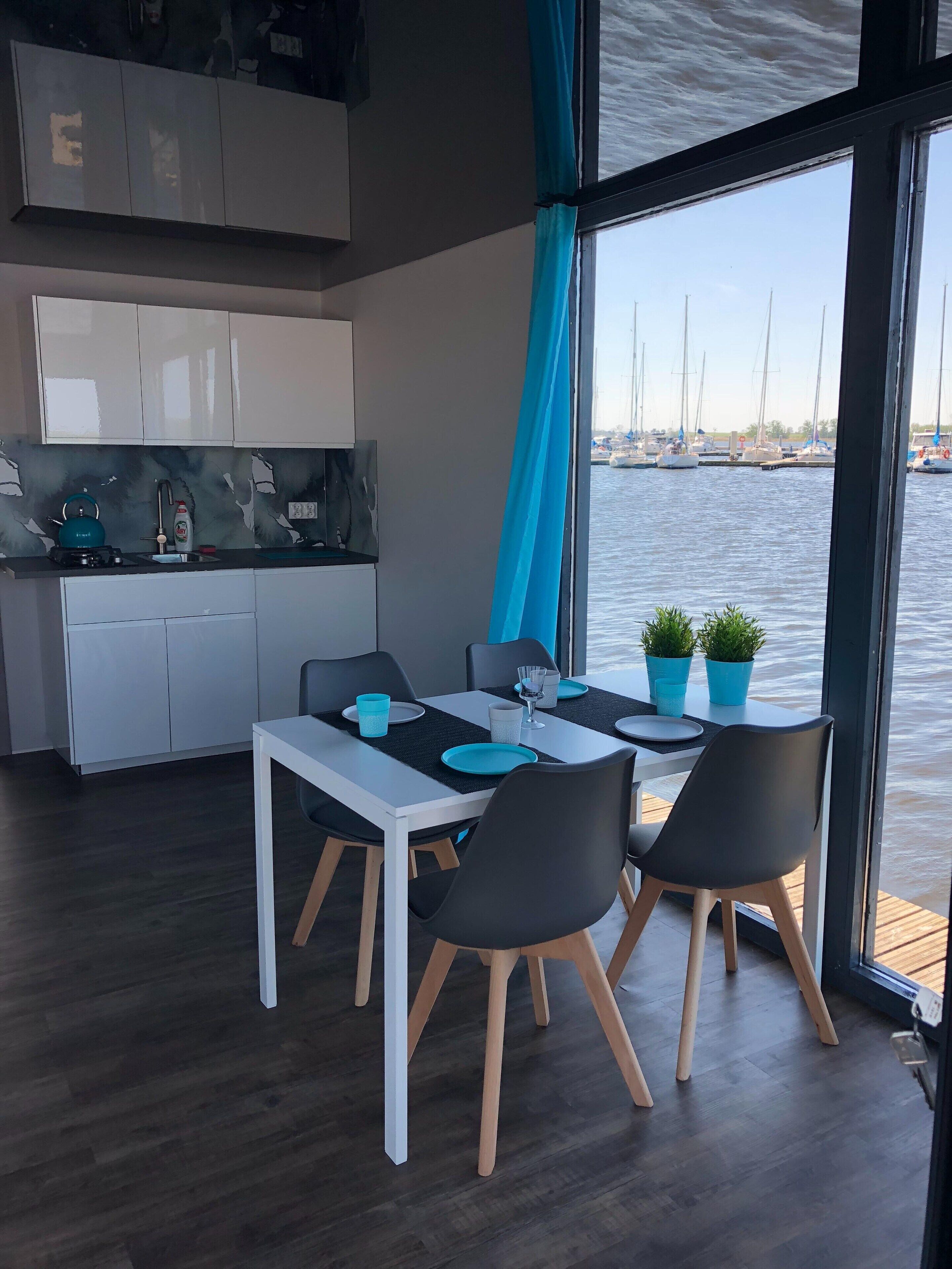 A modern kitchen with white cabinets and a black countertop. There is a dining area with a white table and black chairs. The room has large windows with blue curtains, offering a view of the water and boats outside.