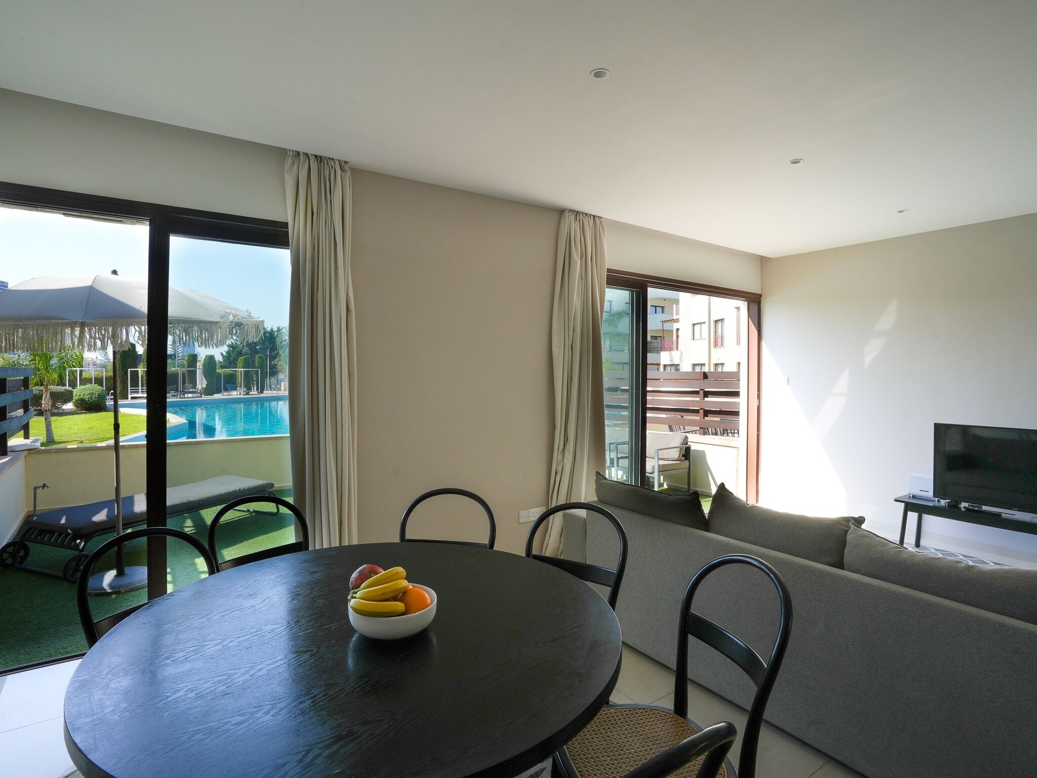 A modern living room with a round table, a sofa, and a TV. A pool is visible through the large windows.