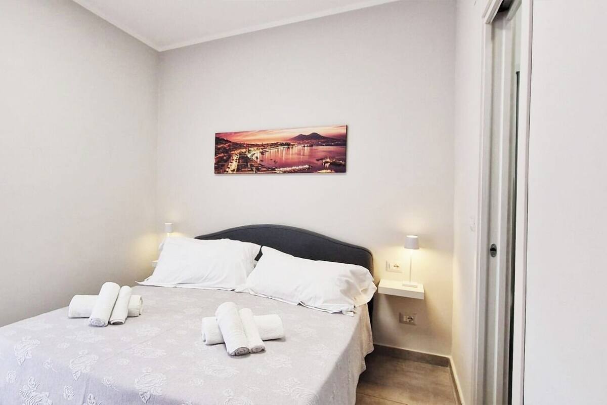 A neatly made bed with rolled towels, a cityscape painting on the wall, and a nightstand with a lamp.