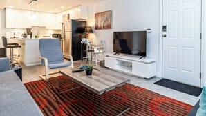 Un salon moderne avec un téléviseur, des sièges, une cuisine à l’arrière-plan et une porte.