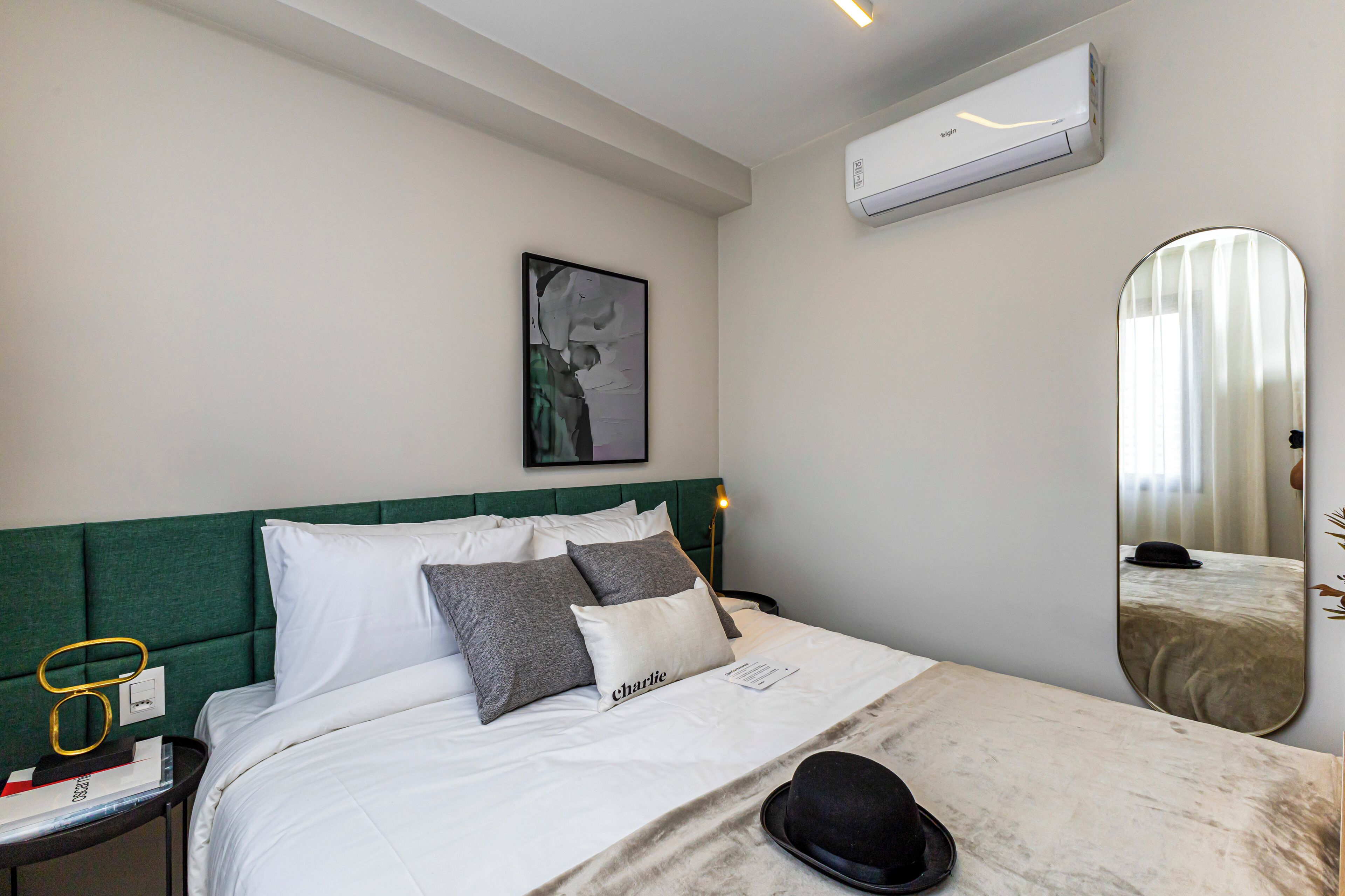 Quarto de hotel com uma cama bem arrumada, uma mesa de cabeceira com um chapéu, um quadro na parede e um aparelho de ar condicionado.