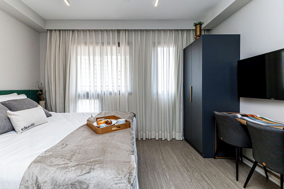 Quarto de hotel moderno com cama bem arrumada, mesa de cabeceira de madeira com itens de café da manhã, escrivaninha com cadeira e uma janela grande com cortinas leves.