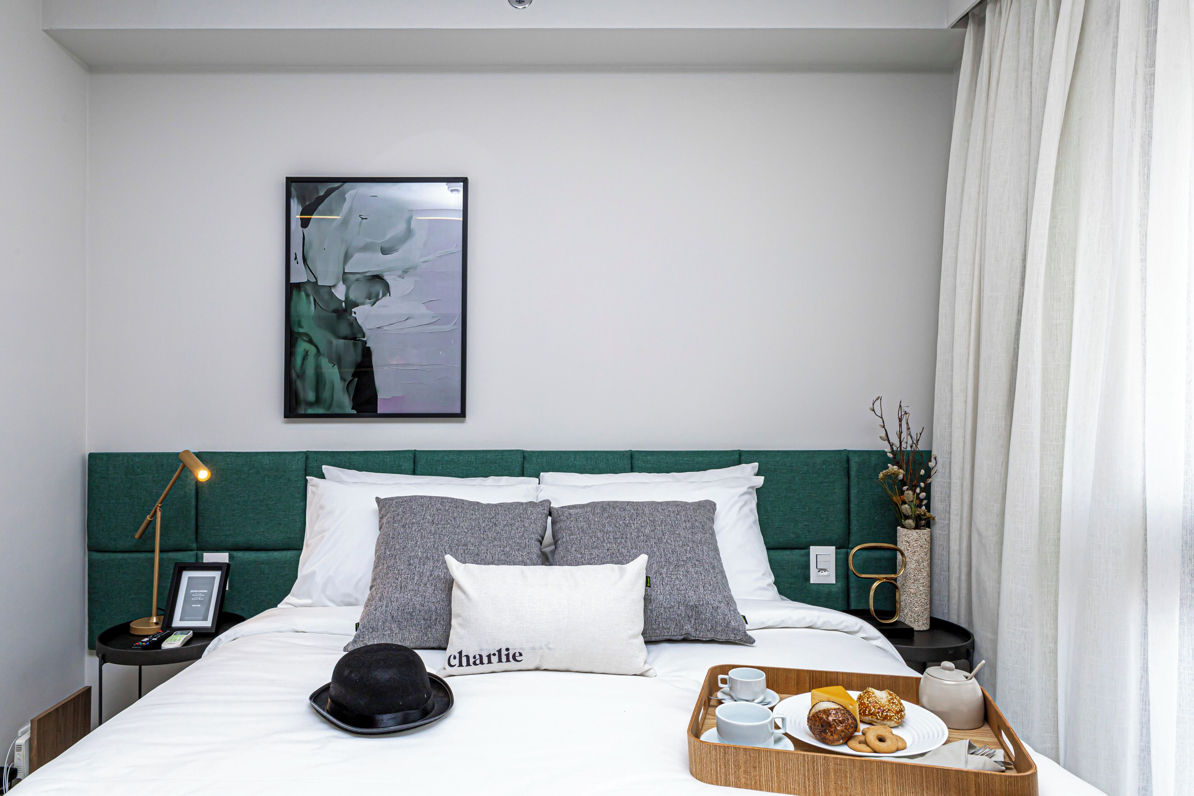 Ein ordentlich bezogenes Bett mit grünem Kopfteil, ein gerahmtes Bild an der Wand und ein Tablett mit Frühstücksangeboten.