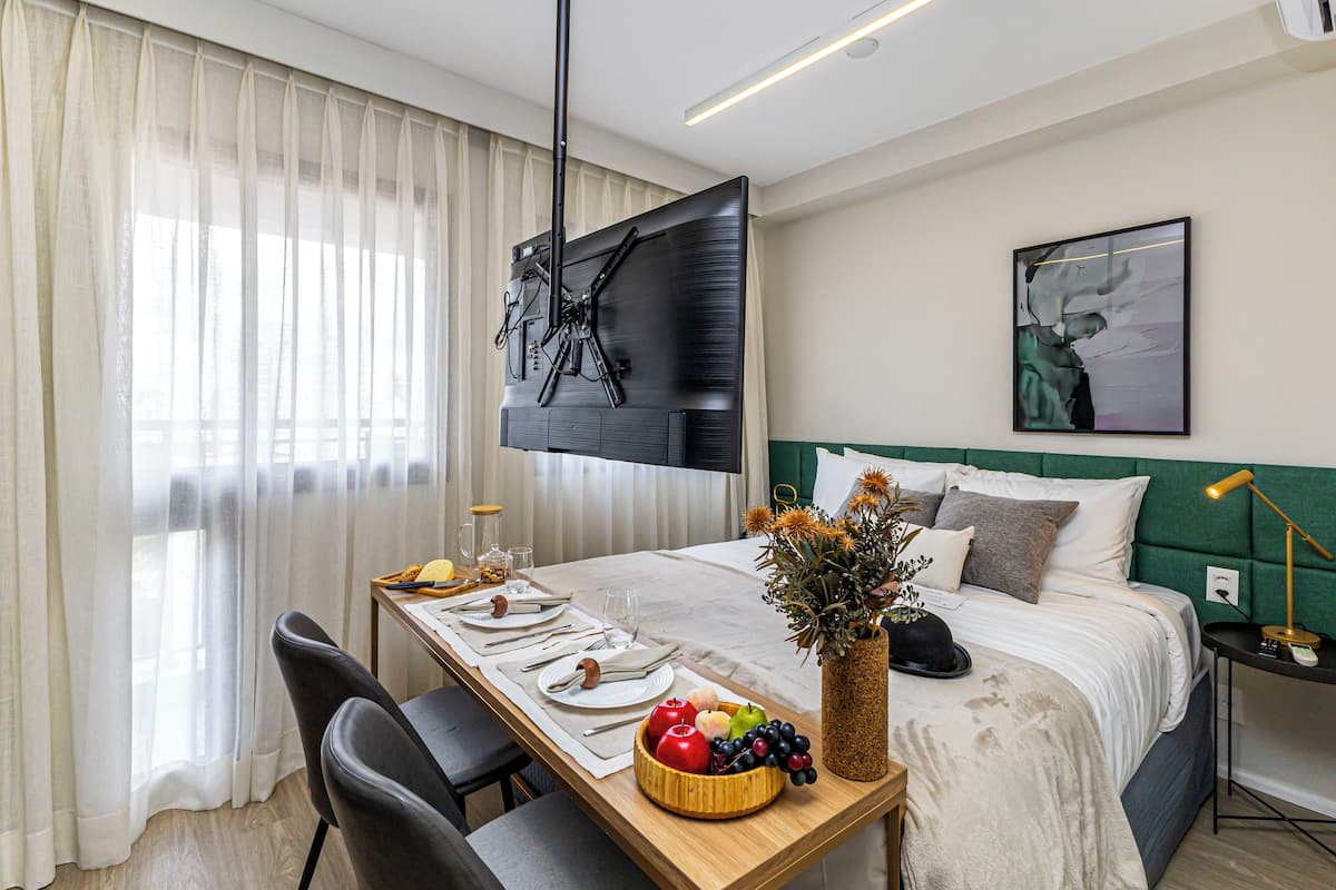 Quarto de hotel bem arrumado, com uma cama, uma mesa de jantar posta para dois, uma televisão na parede e um quadro acima da cama.