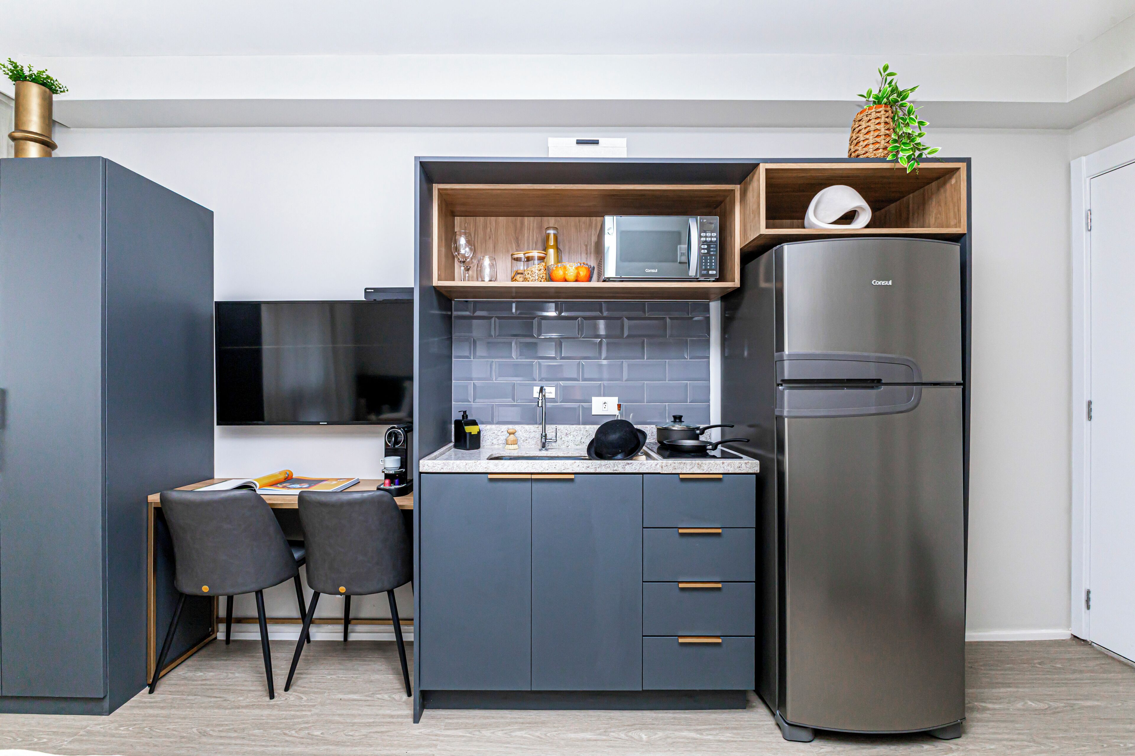 A compact kitchen with grey cabinetry, a stainless steel refrigerator, a microwave, and a wooden shelf with decorative items.