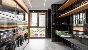A modern laundry room with multiple washing machines, a large window, and a kitchen area with black tiles and wooden shelves. - Charlie Atmosfera Santo Amaro (São Paulo)
