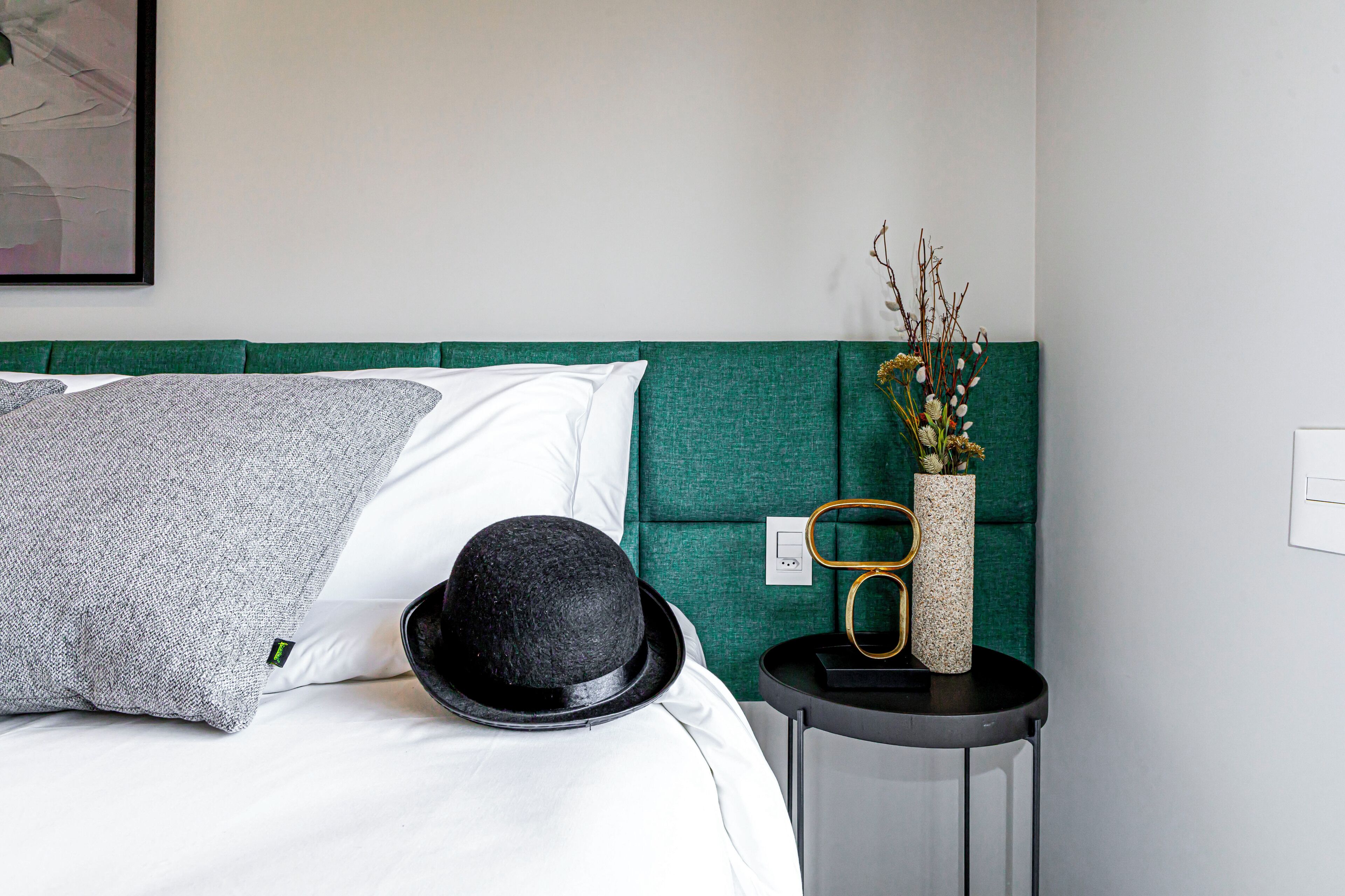 Een net opgemaakt bed met witte en grijze kussens, een zwarte hoed en een vaas met bloemen op een nachtkastje.
