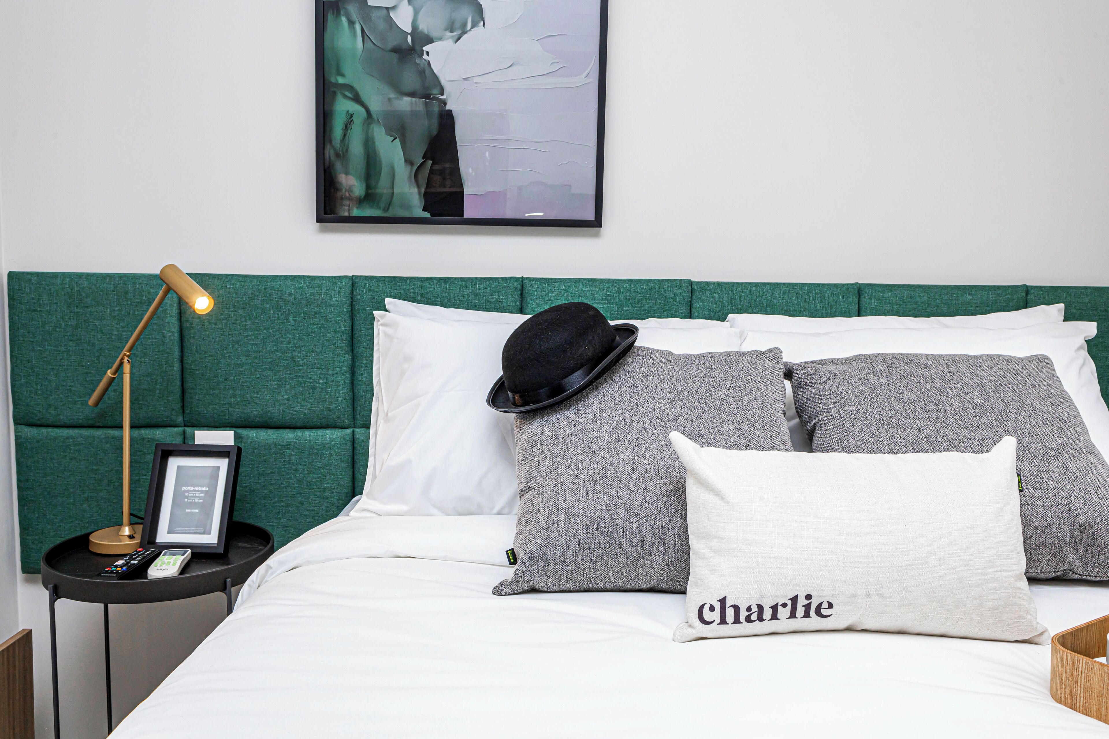 Una cama bien tendida con un cabecero verde, almohadas blancas y grises, un sombrero negro y un control remoto sobre una mesita de noche.