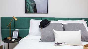Una cama bien tendida con un cabecero verde, almohadas blancas y grises, un sombrero negro y un control remoto sobre una mesita de noche.