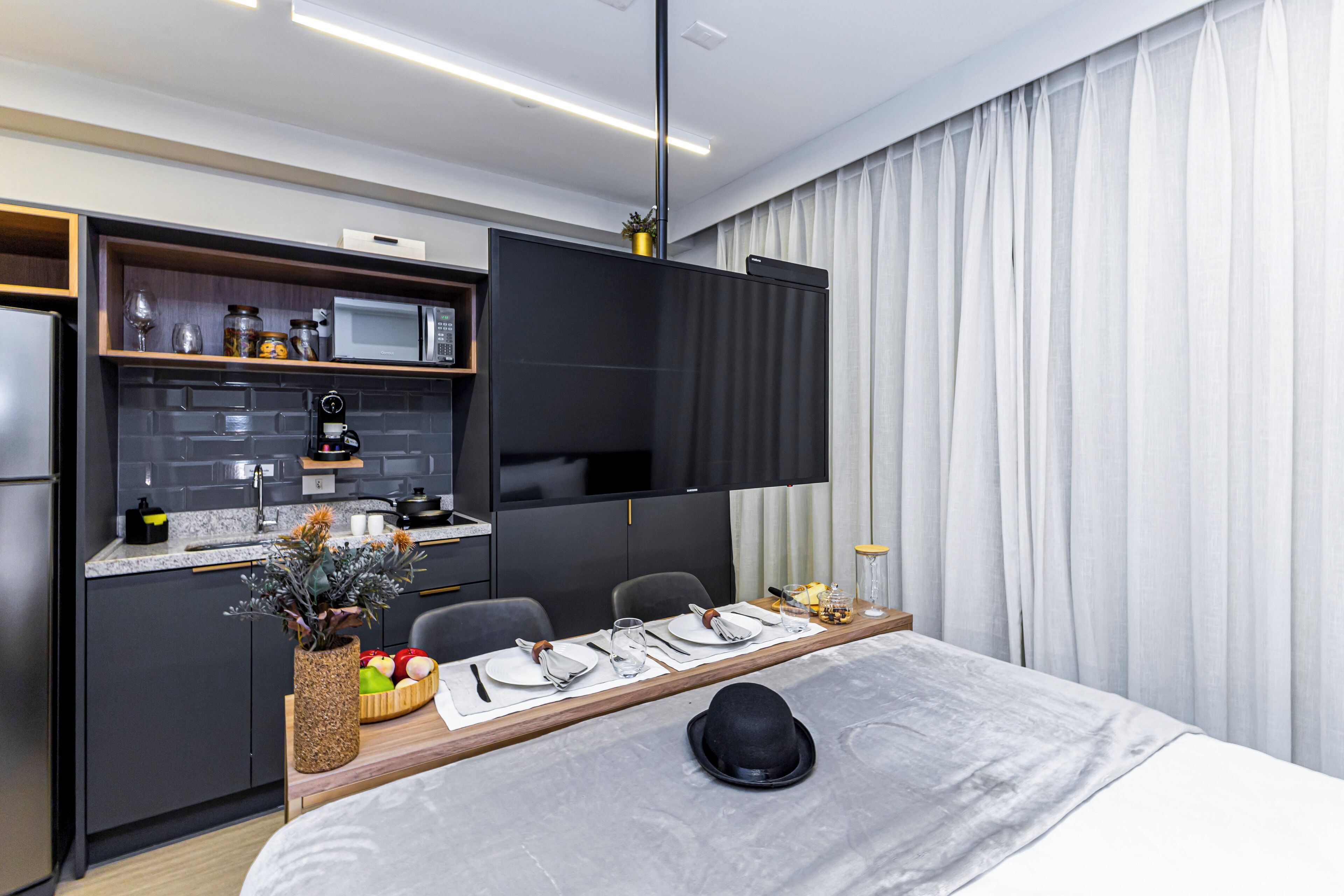 A modern hotel room with a dining area, kitchenette, and a large window with curtains.