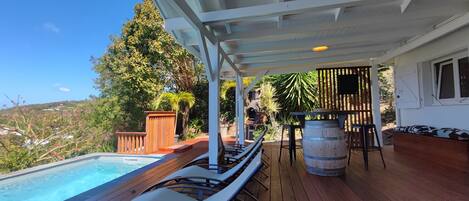 A covered outdoor area with a pool, lounge chairs, a wooden deck, and a bar area.