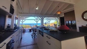 Een ruime keuken met uitzicht op de oceaan, een eetgedeelte met buitenmeubilair en een groot eiland met een rode kom.