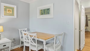 Un comedor con una mesa de madera y sillas blancas, obras de arte en las paredes y un armario blanco con una lámpara.