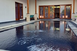 A swimming pool with a clear water surface, surrounded by a building with large glass doors and a stone column. - Villa - Sleeps 6 - Private Pool - Parking (Karangasem)