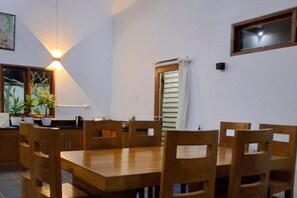A dining area with a wooden table and chairs, a window with blinds, and a wall-mounted light fixture. - Villa - Sleeps 6 - Private Pool - Parking (Karangasem)