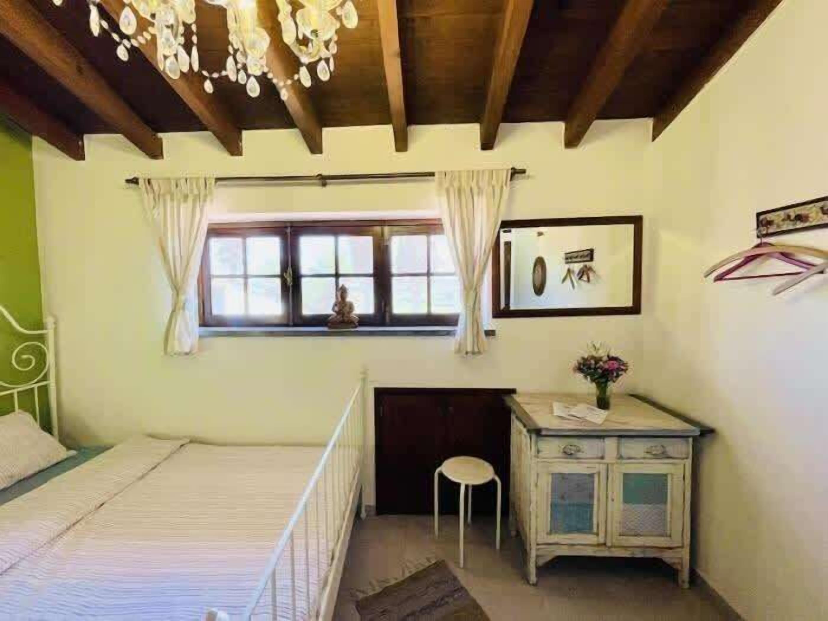 A bedroom with a bed, a desk, a chair, a window with curtains, a hanging light fixture, and a wall-mounted picture.