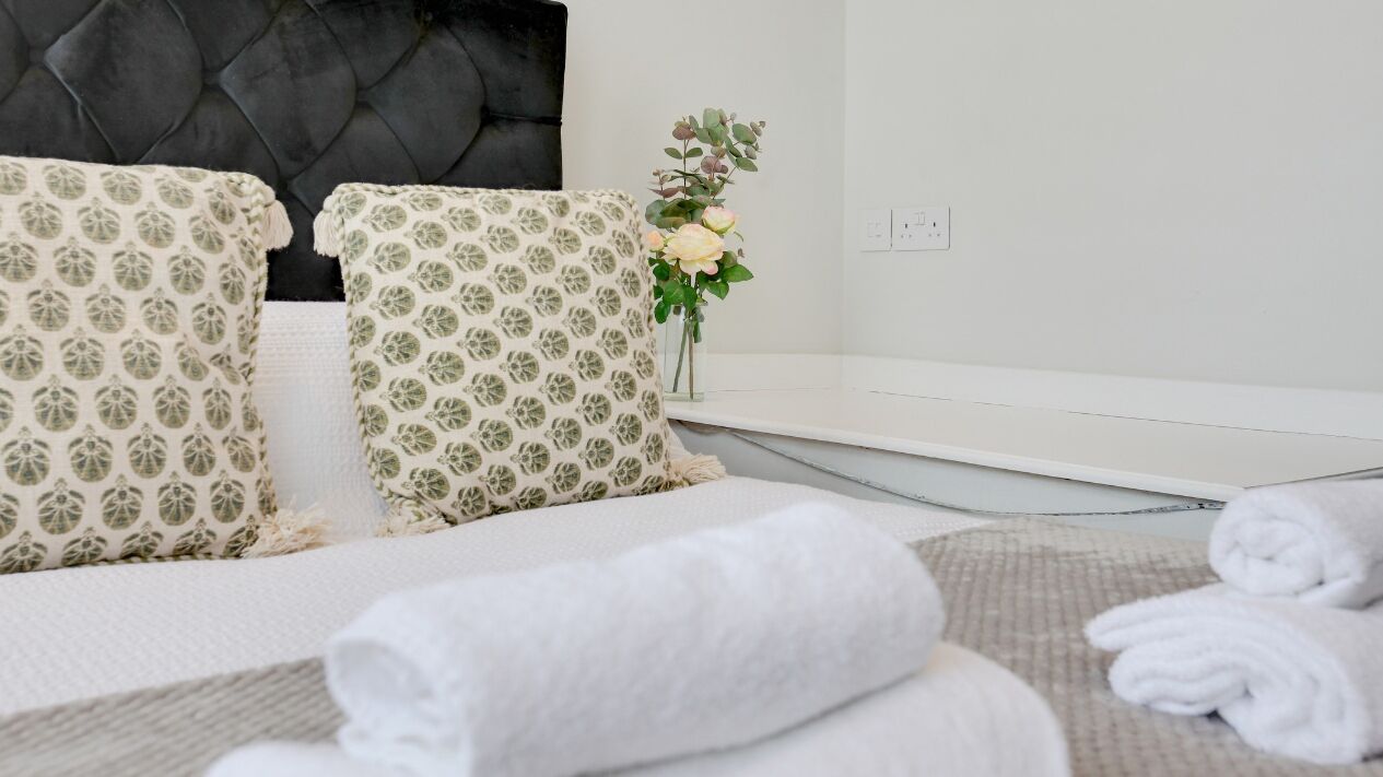Una cama bien hecha con almohadas estampadas, un jarrón con flores y toallas enrolladas sobre una mesita de noche blanca en una habitación de hotel.