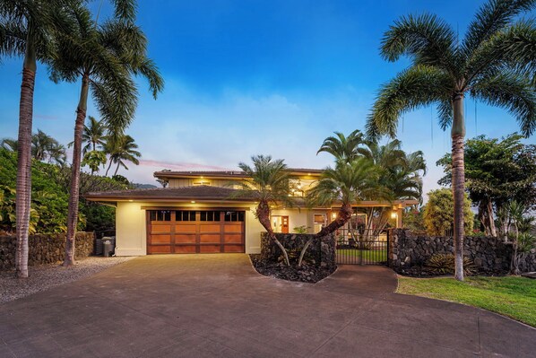 A two-story house with a garage, surrounded by palm trees and a stone wall.