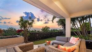 A covered outdoor seating area with wicker furniture, a stone wall, and a view of the sunset.