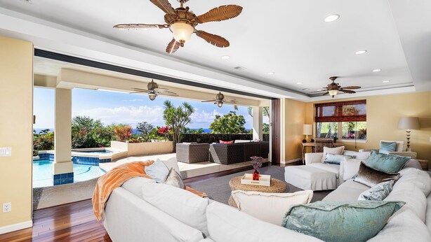 A spacious living area with a large white sofa, a coffee table, and a pool view. There are ceiling fans and recessed lighting.