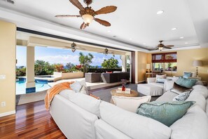 A spacious living area with a large white sofa, a coffee table, and a pool view. There are ceiling fans and recessed lighting.