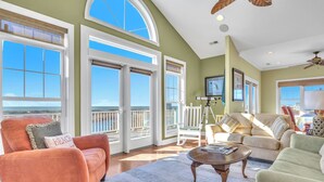 A spacious living room with large windows, a ceiling fan, and a view of the ocean. - The Loggerhead: Oceanfront, Beautiful Walkway Over Marsh (Oak Island)