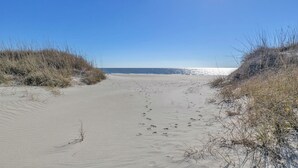 A sandy beach with footprints leading to the water, surrounded by dunes. - The Loggerhead: Oceanfront, Beautiful Walkway Over Marsh (Oak Island)