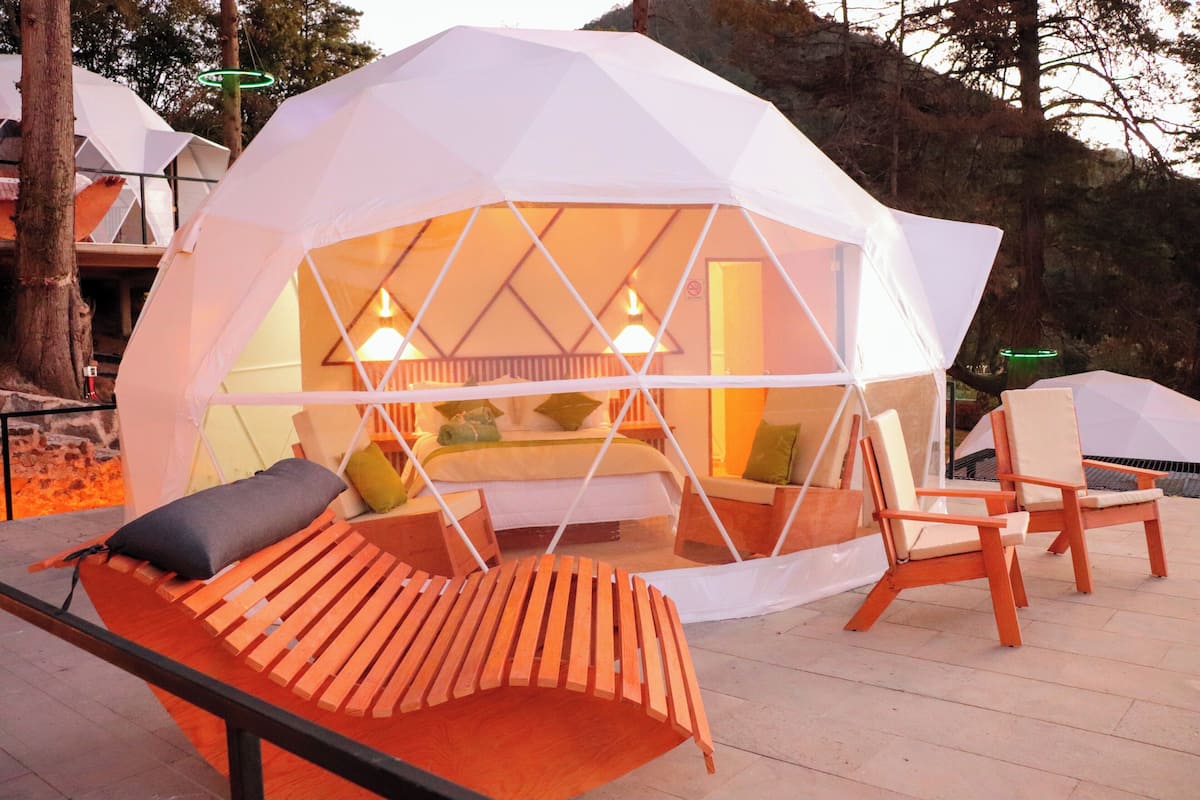A geodesic dome structure with a bed and seating area inside, surrounded by trees.