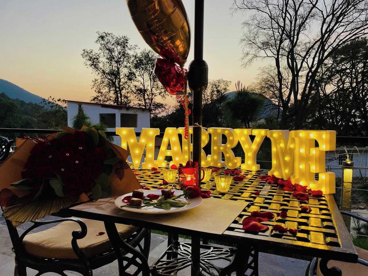 Une table dressée pour un mariage, avec des lettres lumineuses formant les mots « ÉPOUSE-MOI » et un bouquet de roses rouges.