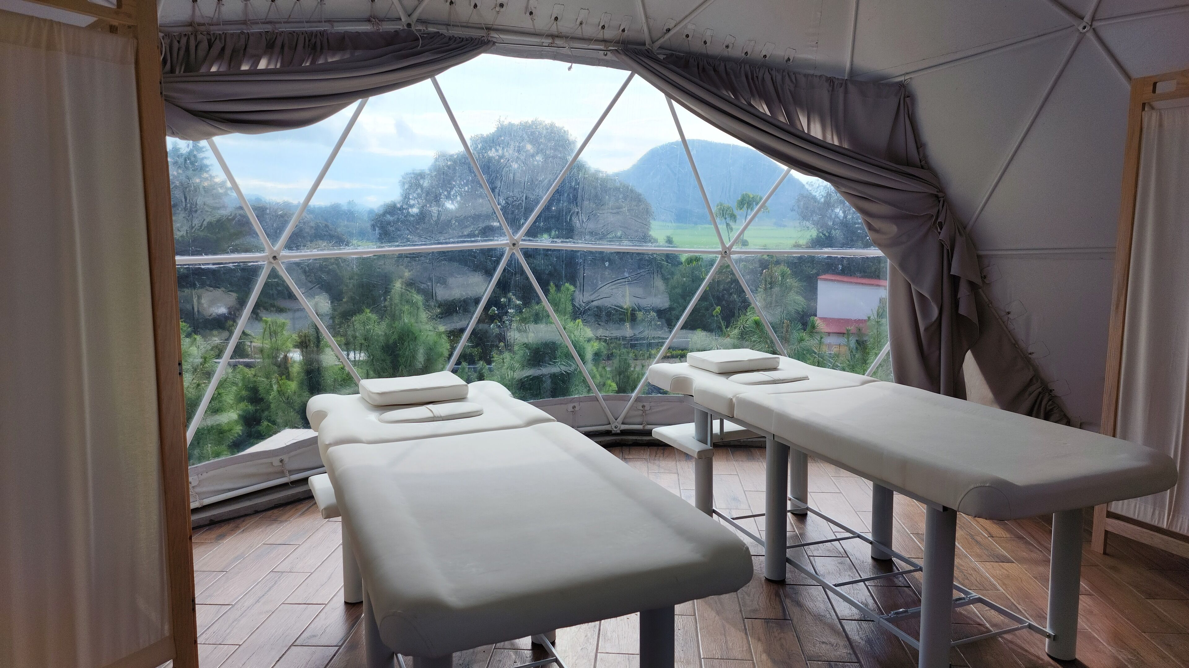 A room with two treatment beds, a large window with a view of greenery and mountains, and a ceiling with a geometric pattern.