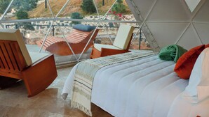 A room with a bed, two wooden chairs, a view of a hillside through a glass dome structure.