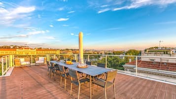 Eine Dachterrasse mit Tisch und Stühlen, einer Flagge und Blick auf Gebäude und Bäume.