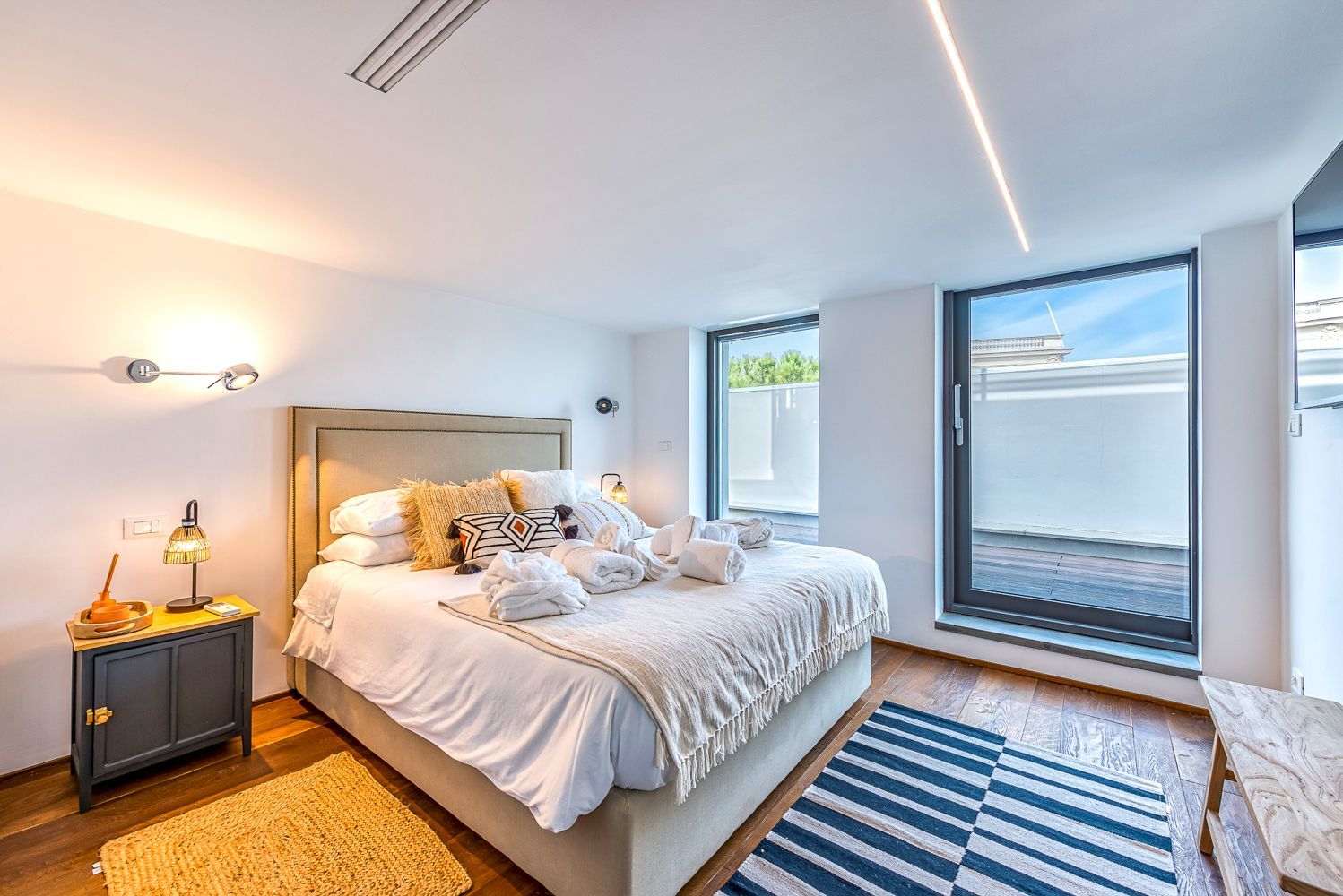 A modern bedroom with a large bed, bedside lamps, a nightstand, and a view of the outdoors through a large window.
