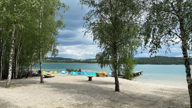Järvenrantainen alue, jossa on veneitä, puinen penkki ja hiekkaranta, jota koivupuut ympäröivät.