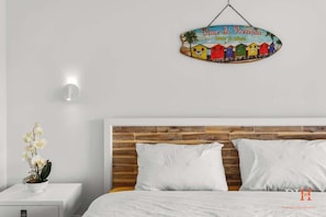 A bedroom with a wooden headboard, white bedding, a small bedside table with a plant, and a wall-mounted sign above the bed. - Amazing Location with Ocean View (Cowes)