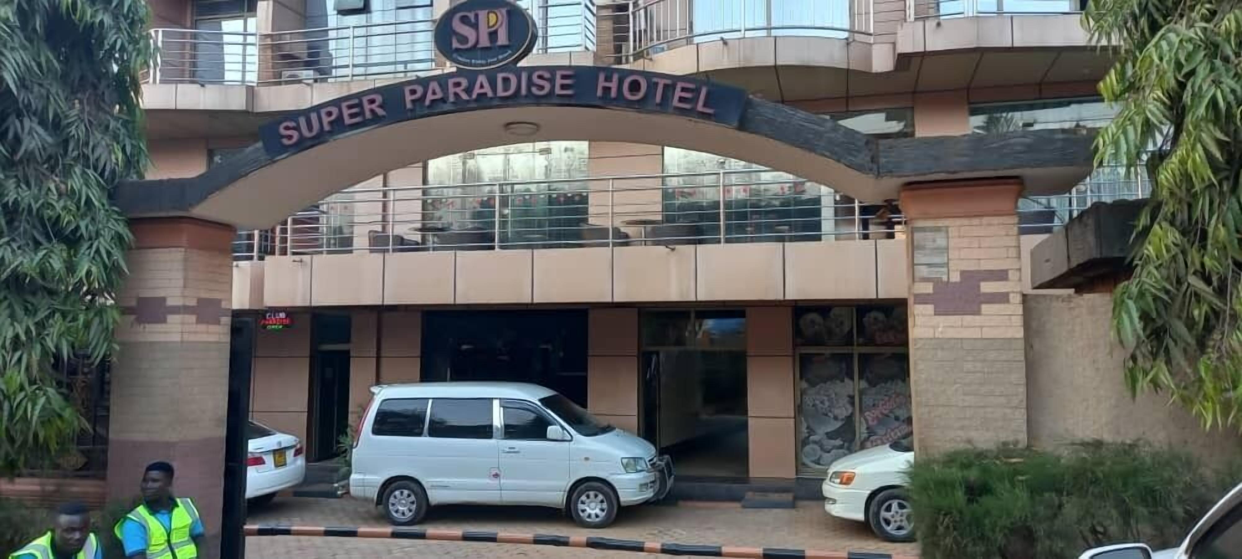 The entrance to Super Paradise Hotel with a clear sign, a white car parked in front, and a security guard standing nearby.