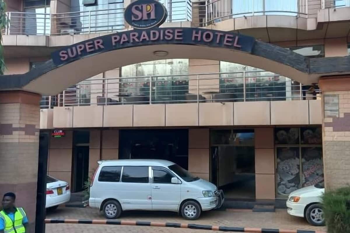 La entrada del Hotel Super Paradise, con un letrero visible, un coche blanco estacionado adelante y un guardia de seguridad cerca.
