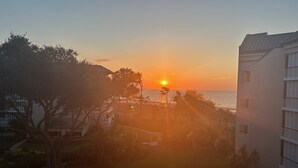 Une vue sur le coucher de soleil depuis le balcon d’un hôtel, donnant sur une plage bordée de palmiers et un ciel dégagé.