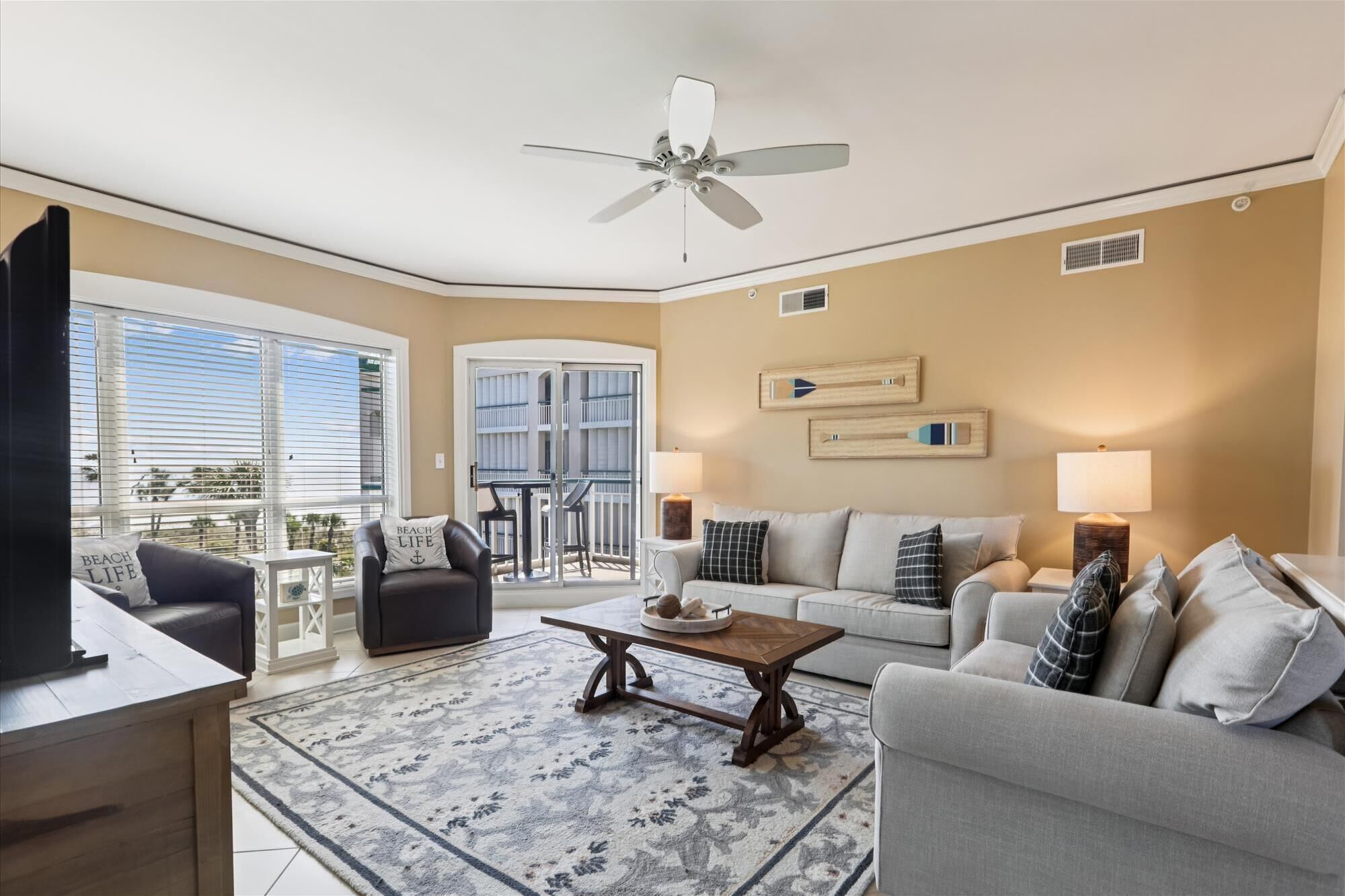 A spacious living room with a ceiling fan, a large window with blinds, a balcony, and a dining area with a table and chairs.
