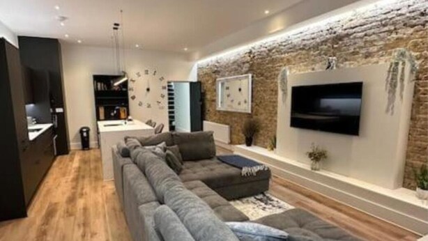 Een moderne woonkamer met een grote grijze hoekbank, een flatscreen televisie aan een bakstenen muur en een houten vloer.