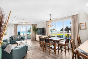 A spacious living area with a dining table, chairs, a sofa, a TV, and large windows offering a view of the beach.