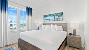 Un dormitorio con una cama grande, una mesita de noche con lámpara, una ventana con cortinas y un cuadro de una playa.