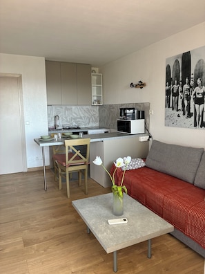Cocina moderna con microondas, tostadora y cafetera. Cuenta con una zona de comedor con mesa y sillas, y un salón con sofá y un florero.