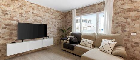 A living room with a stone wall, a flat-screen TV on a white cabinet, a beige sofa, and a window with curtains.