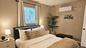 Een slaapkamer met een bed, nachtkastje, lamp, potplant en een bord aan de muur.