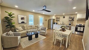 Een ruime woonkamer met een bank, fauteuil, salontafel, eettafel en stoelen. Er is een plafondventilator, een keuken met apparatuur, en een groot raam.