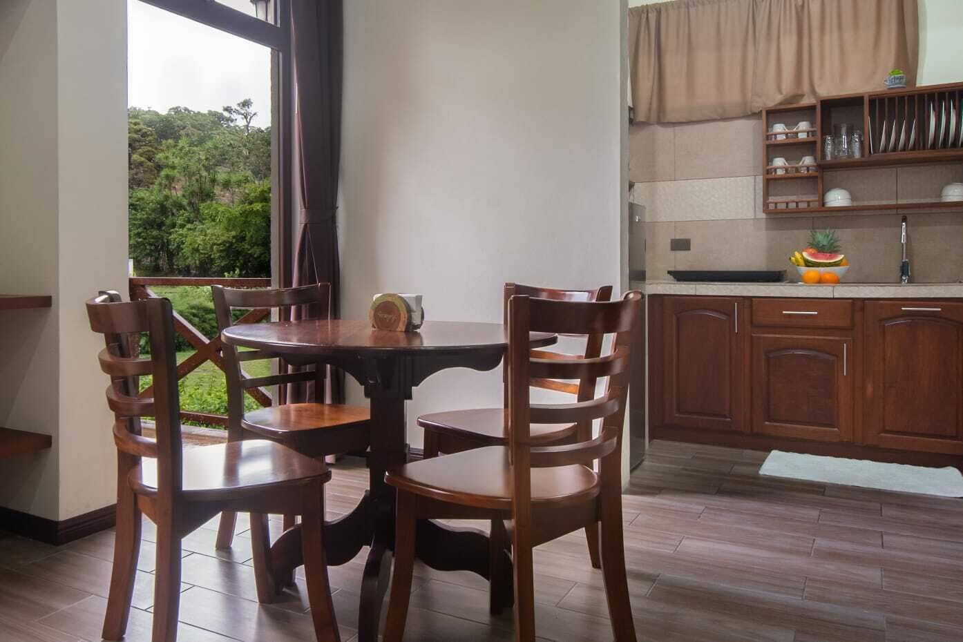 Un coin repas avec une table ronde et quatre chaises, une cuisine dotée de meubles en bois et d’un panier de fruits, et une vue sur la verdure par une fenêtre.