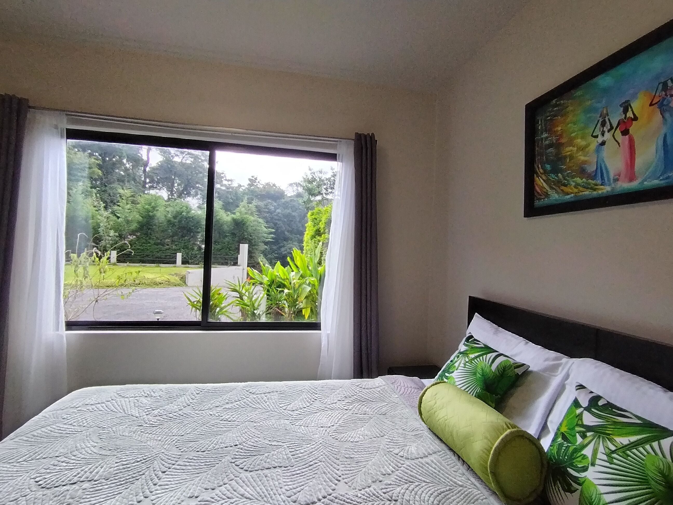 A hotel room with a large window, a bed with a white textured bedspread, green pillows, and a painting on the wall.
